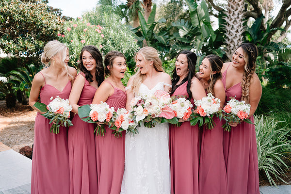 Bride and Bridesmaids gathered together, holding flower bouquets, in front of a tropical background with lots of greenery and palm trees.