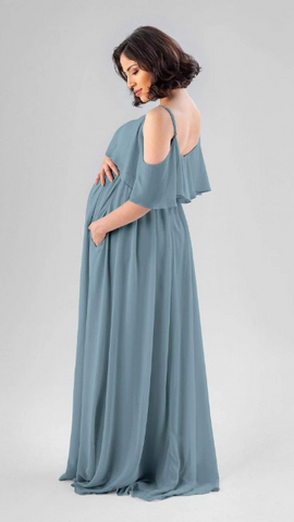Kennedy Blue model wearing a maternity dress with a ruffled swooped neckline and flowy skirt.