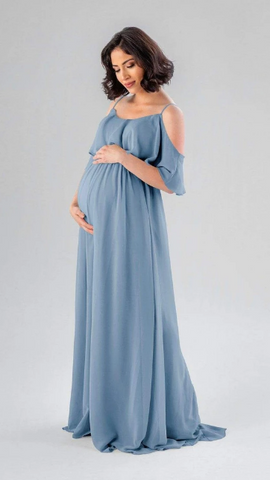 Kennedy Blue model wearing a maternity dress with a ruffled swooped neckline and flowy skirt.