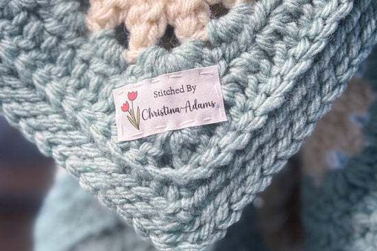 A close-up photo of a hand-crocheted blanket in shades of blue and cream, highlighting a custom cotton fabric label that reads 
