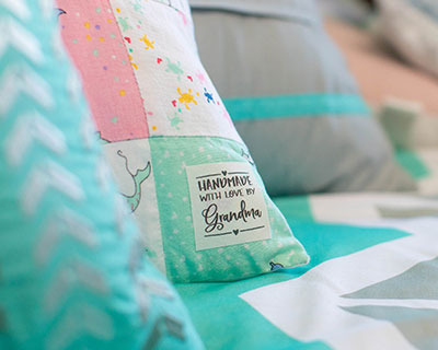 IRON-ON LABELS 101: Quick and Easy Labelling' features a close-up of a quilt with a personalized iron-on fabric label that reads 'handmade with love by Grandma.' The image highlights the ease of using custom iron on labels and iro