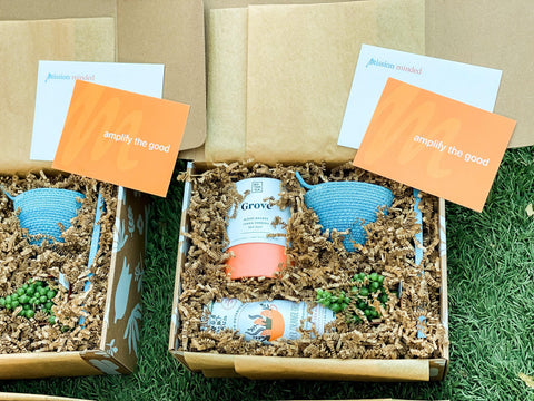 Two gift boxes side by side with Mission Minded notecards featuring tagline "Amplify the Good" and blue and orange colored gifts including a candle, succulent, woven planter and tea tin