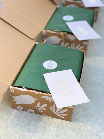 Two kraft gift boxes with a botanical design in white on the exterior are filled with tissue paper enclosure and The Product Boss branded sticker and notecard