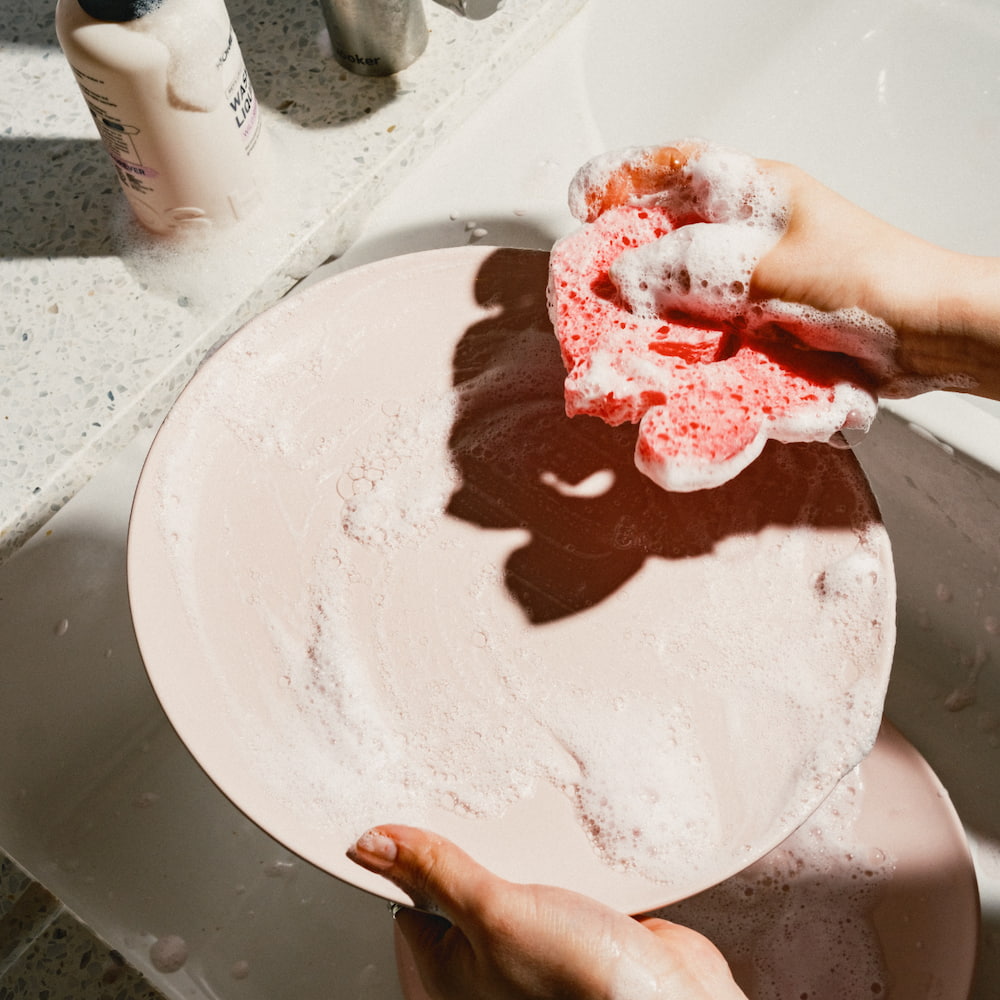 Hand washing a pink plate with soapy sponge in a sunlit sink.