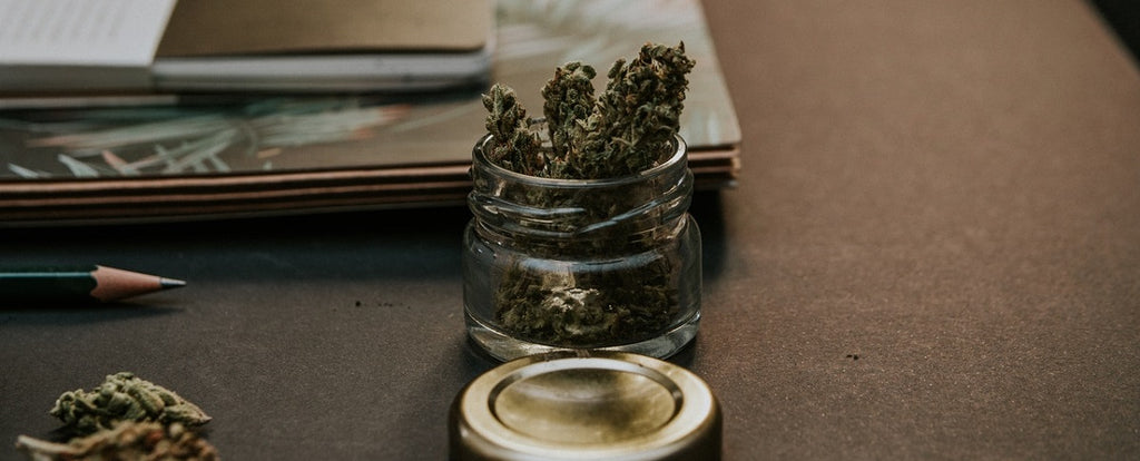 A jar of cannabis on a desk next to a book and pencil.