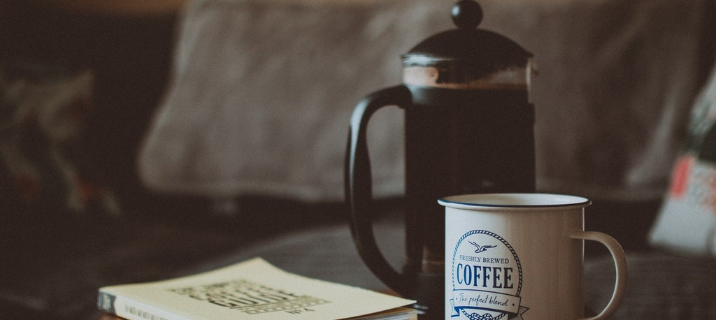 A coffee press beside a book and a mug on a desk.