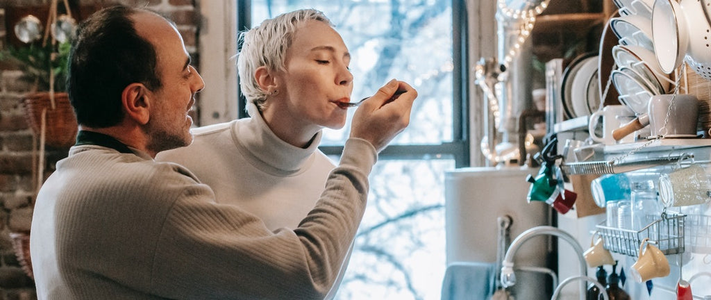 One person offering another person food to taste from a spoon, in front of a dishrack and window.