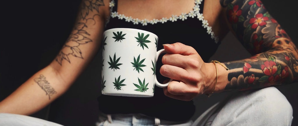 A person with tattoos holding a mug of tea that has cannabis leaves on it.