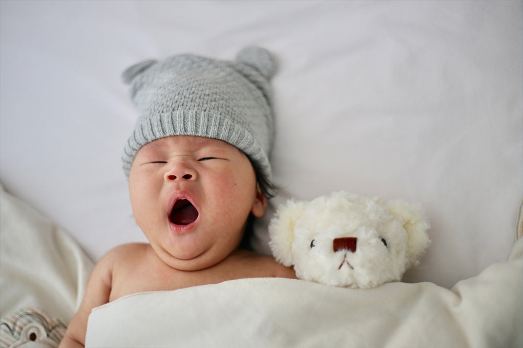 Foto de bebé bostezando acostado en carriola o cuna con un peluche.