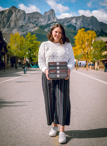 Cofounder Alessa Amato standing in the street in downtown Canmore in front of mountains, holding 4 boxes of Algi's IMPACT Bars