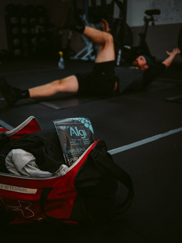 Man at cross fit gym doing core exercises during a workout. In front of him is a vegan protein powder, spirulina algae