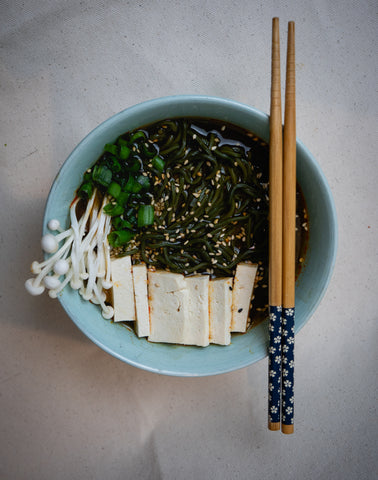 Blue-green bowl of delicious spirulina algae ramen noodles make with vegetable broth, oyster mushrooms, and tofu, topped with sesame seeds and green onions. Placed on top of the blue-green bowl is a pair of chopsticks resting together.