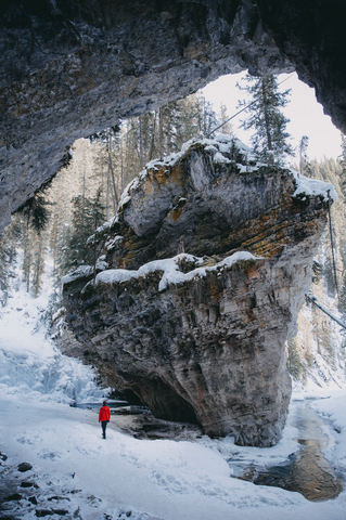 Man in red winter jacket standing at base of giant rock formation in Johnston Canyon Trail. Snow covers the mountains, cliff walls, and trees