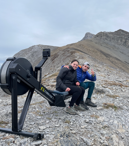 Devon and Alessa sitting on an erg rowing exercise machine on top of Ha Ling mountain in Canmore Alberta