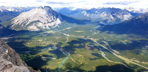 View from the summit of Mount Rundle overlooking the valley of green trees, and blue rivers. A chain of snow capped mountains are seen in the distance