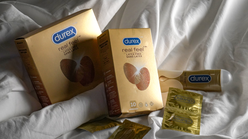 Des condoms naturels sans latex Durex Real Feel et du lubrifiant Durex Real Feel sur un lit aux draps blancs. 