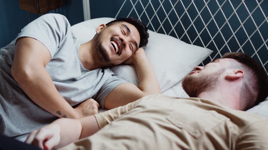 Two men smiling while laying in bed together after practising how to cum with a condom on.