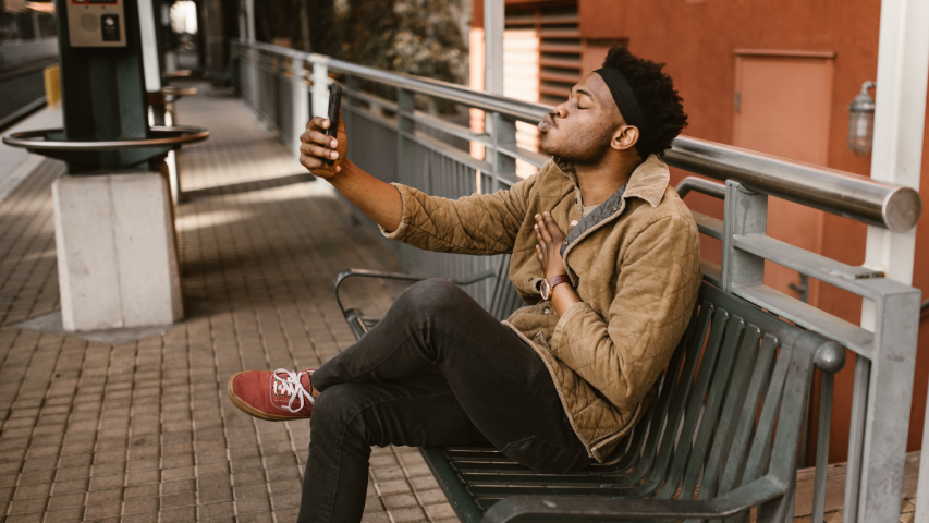 Man sitting on a public bench blowing a kiss to his long-distance partner through the phone.