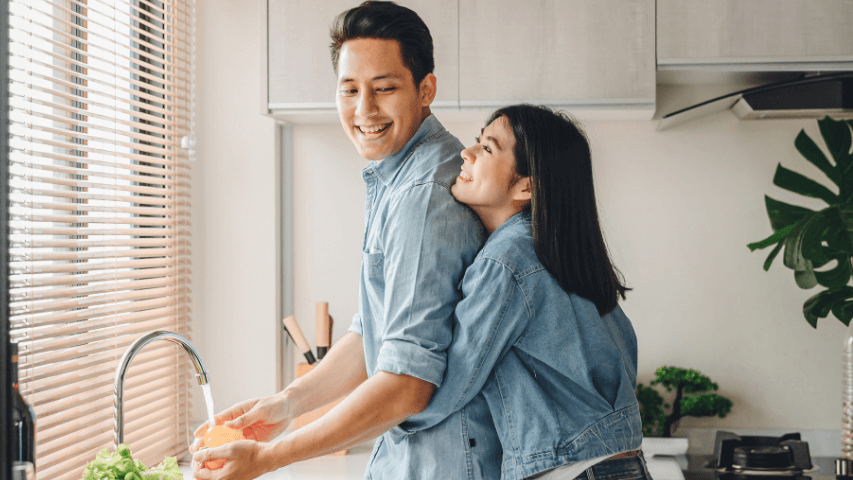 Couple wearing denim jackets smiling and hugging while doing dishes in the kitchen.