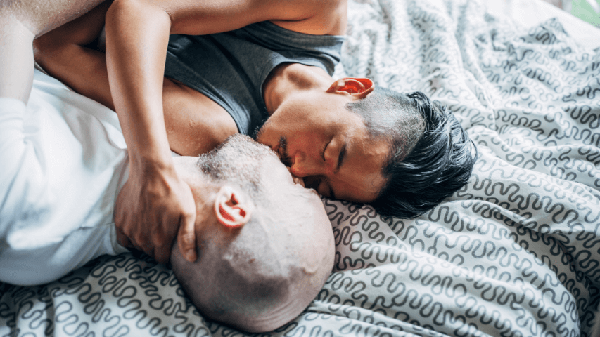 Two men sharing a soft and intimate kiss on their bedspread.