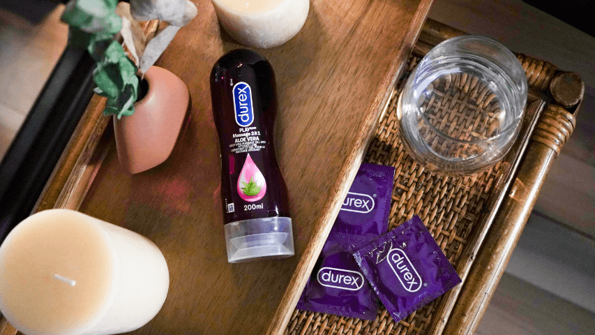 Une bouteille de Play Massage 2 en 1 Aloe Vera de Durex sur une table de chevet avec un tiroir ouvert dans lequel on voit des condoms Durex violets.