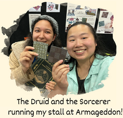 a photo of two of my best friends running a convention stall for me at Auckland Armageddon 2023. Behold the faces of two amazingly wonderful and kind people! <3