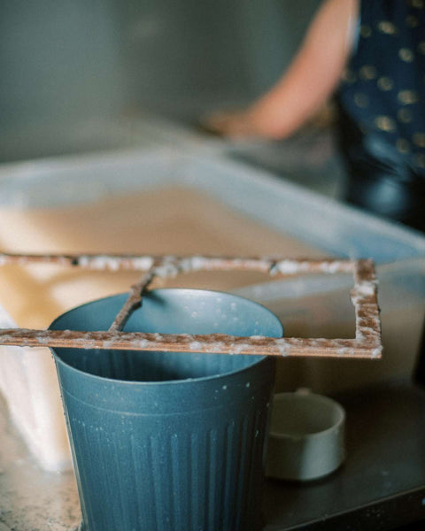 handmade paper making wood frame speckled with cotton bits and a woman working in the backgorund 