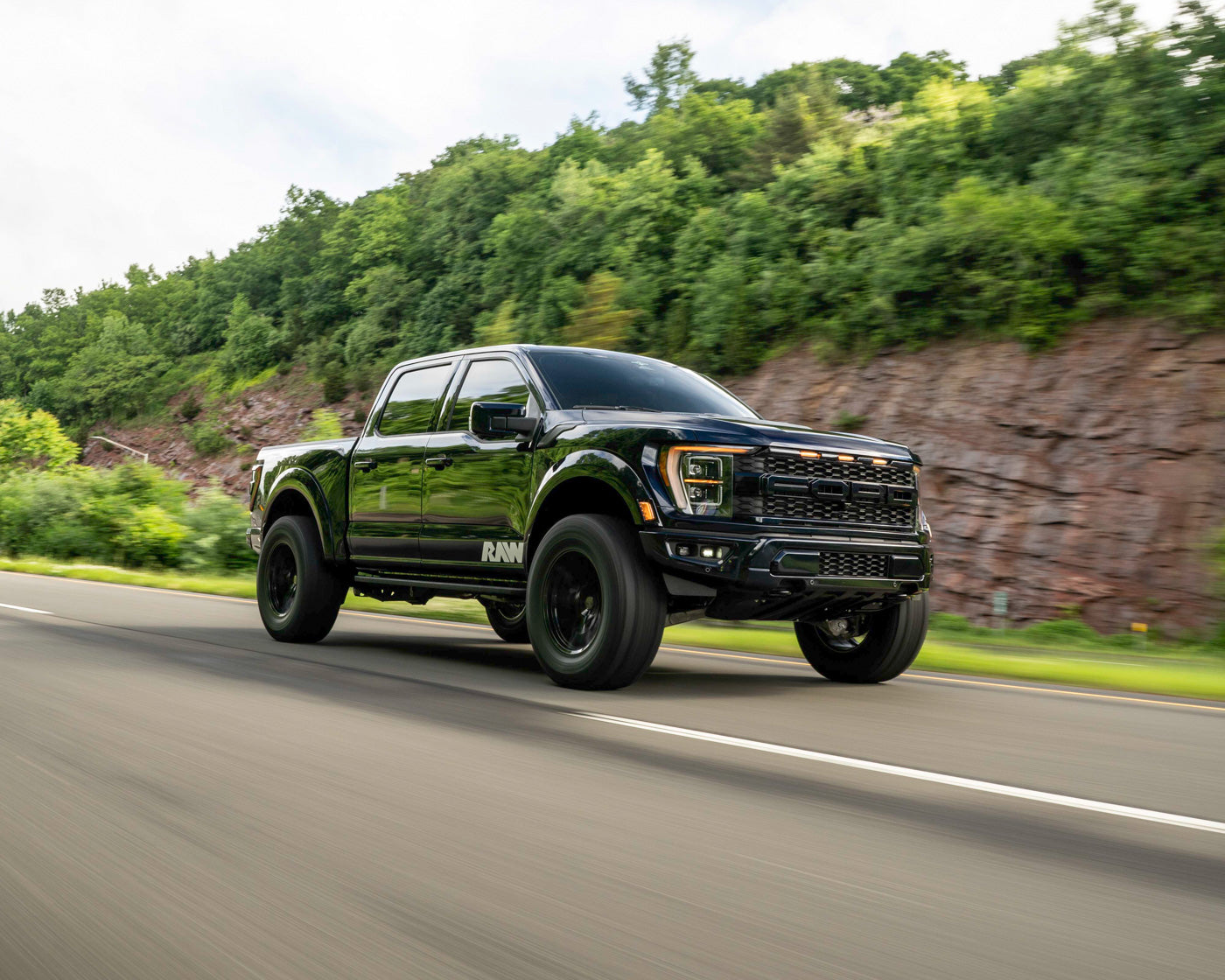 Black pickup truck driving on a road with a green hillside in the background.