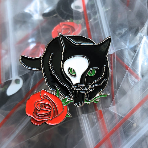 A soft enamel pin of a black cat wearing a half-mask and holding a rose, by Braden Duncan