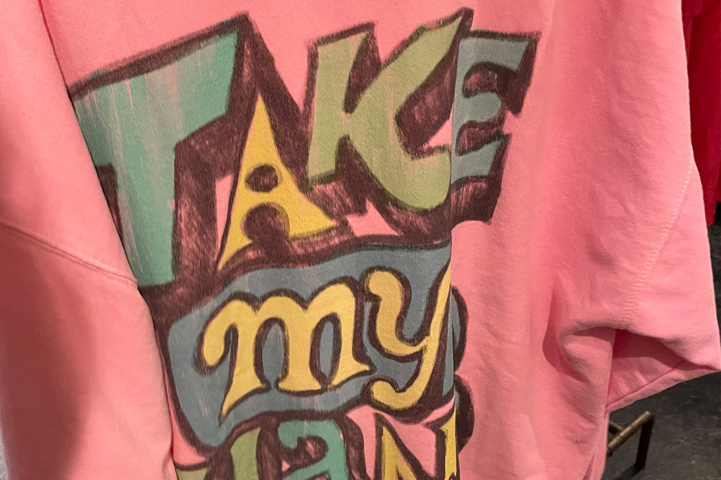An oversized pink t-shirt that has the hand drawn text ‘Take My Hand’ covering the back.