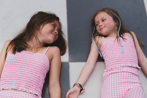 Two teen girls are laying down side by side wearing matching Pink Gingham Shirred Strappy Girls Crop Top and Pink Gingham Print Girls Leggings  from Gen Woo’s Retro Psychedelia Spring fashion collection.