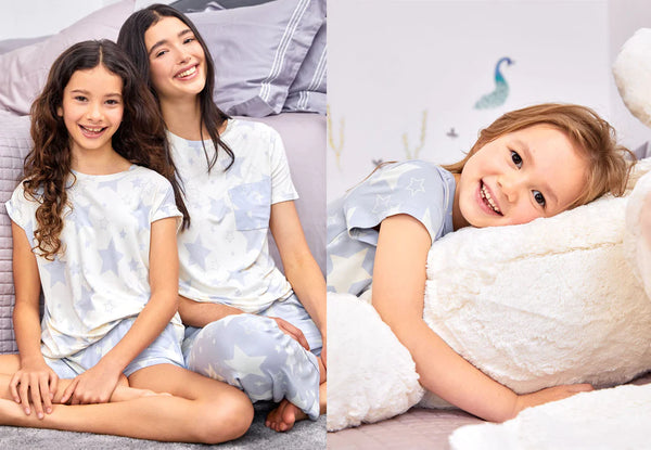 A happy mum and daughter wearing matching star print pyjamas, sitting beside a bed and smiling at the camera.