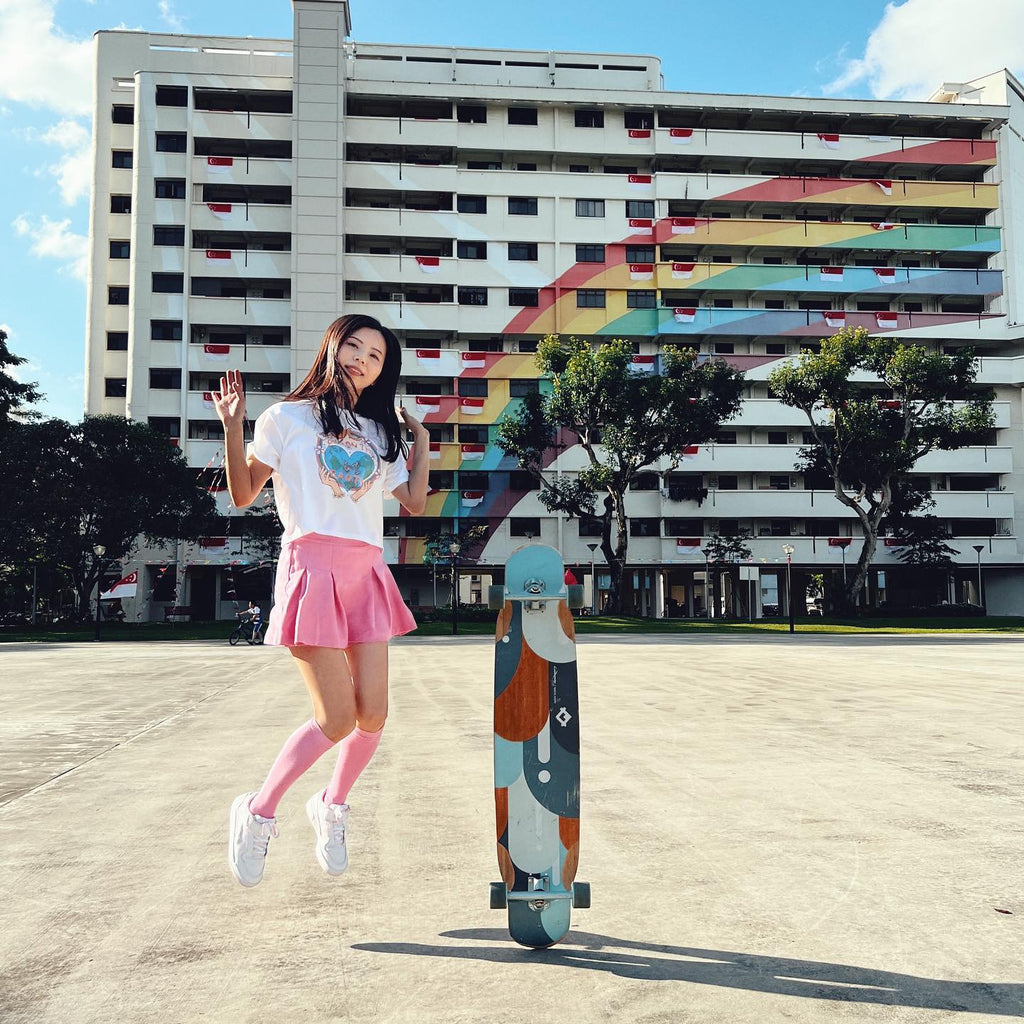 Confident longboard skater jumps up by her board, in colourful skate wear, in front of a rainbow building.