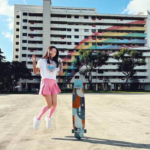 Longboarder Lucy jumps up in the air in front of a building decorated with a rainbow in Singapore. She wears a pink skort by Gen Woo.