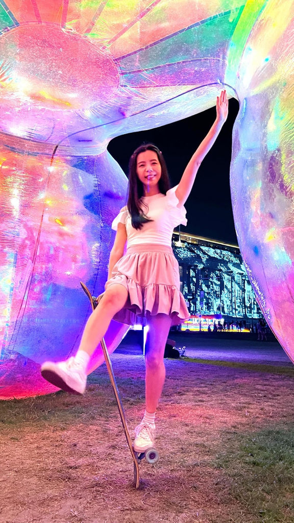 A happy skater stands on her longboard in front of the bright city light show, smiling into the camera.