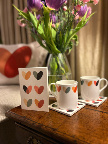 Image of tulips in a vase with a throw cushion in the background with a red and orange heart print on it. Mugs and a card in the foreground with colourful heart print. From Mustard and Gray