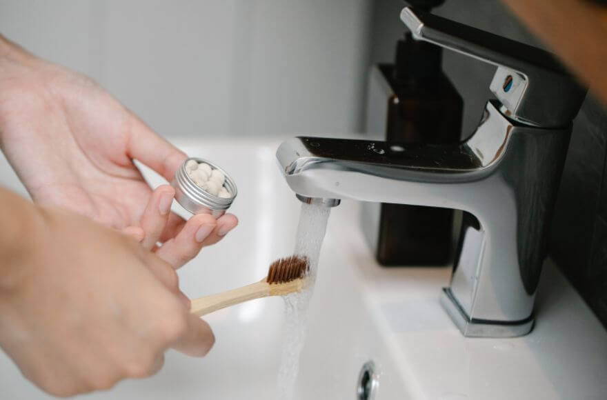 Woman holding a bamboo toothbrush under a tap, with eco toothpaste tabs in the other hand.