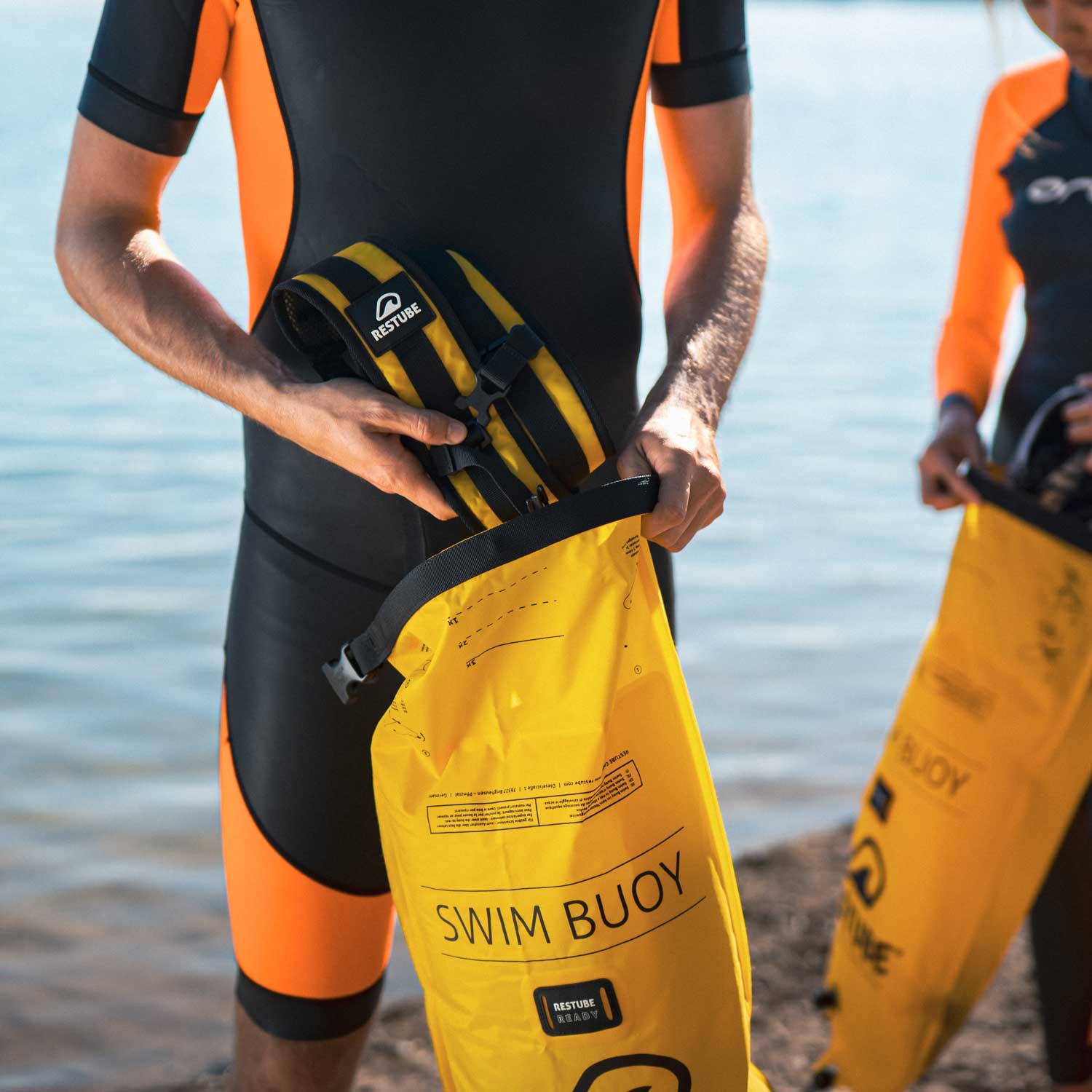 man standing at the lake holding the swimbag and putting the backpack straps into the bag