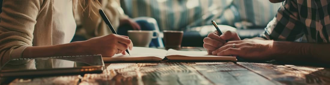 A couple sitting at a table, journal open in front of them, sharing a pen as they write down their favorite shared memories.