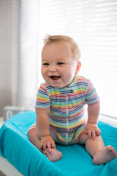 smiling baby wearing a rainbow stripe shortie, sitting on a changing table