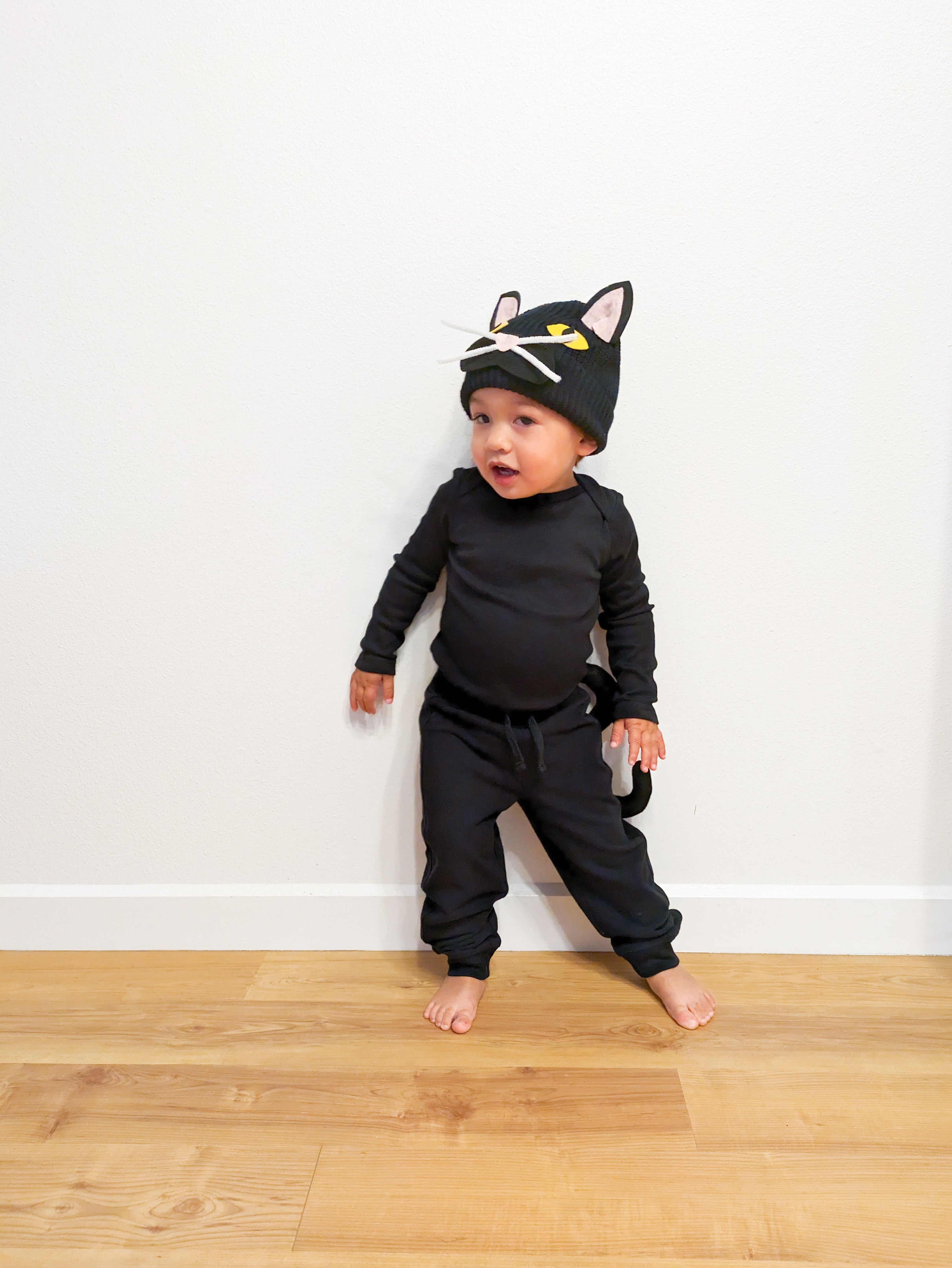 A toddler dressed as a black cat, in a black romper with DIY ears and whiskers attached to a black hat