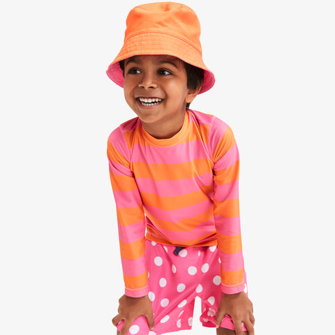 Young boy smiling in an orange sun hat, a long sleeve rash guard with pink and orange stripes, and pink and white polka dot swim trunks