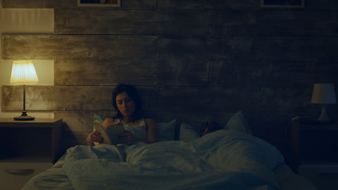 Woman reading physical book with nightstand lamp on while husband sleeps