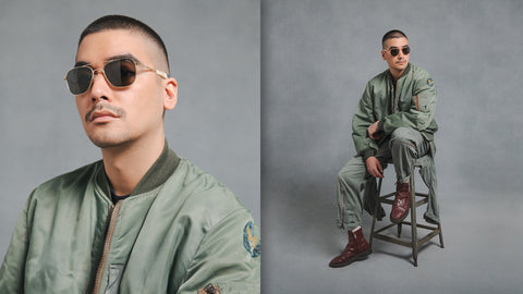 A young man wearing military streetwear and aviators sits on a stool in front of a grey background