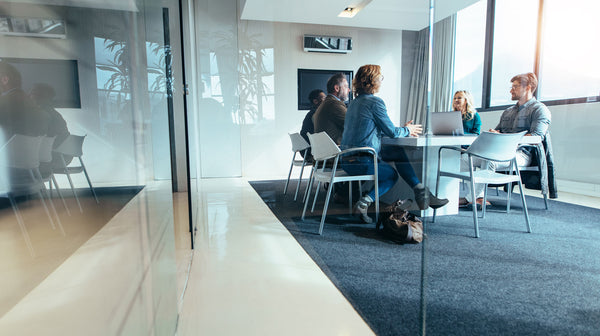 Through the glass walls of a contemporary meeting room, a group of professionals is engaged in a discussion around a minimalist white table, featuring ergonomic office chairs and a clean, open office environment with natural light.