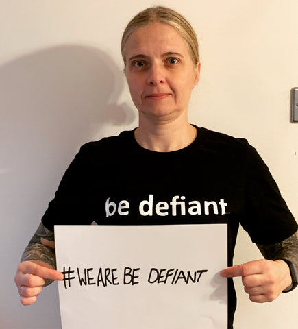 woman with tattooed arms in a black shirt holding a sign saying '#WeAreBeDefiant