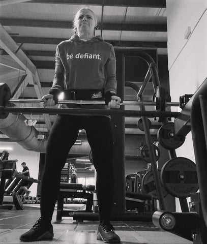 Black and white picture of a woman standing up doing bicep curls in the gym in a be defiant hoodie