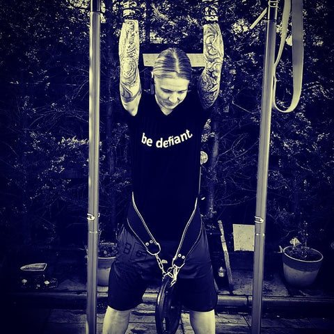 woman with tattooed arms doing pull ups outside in a be defiant tshirt smiling