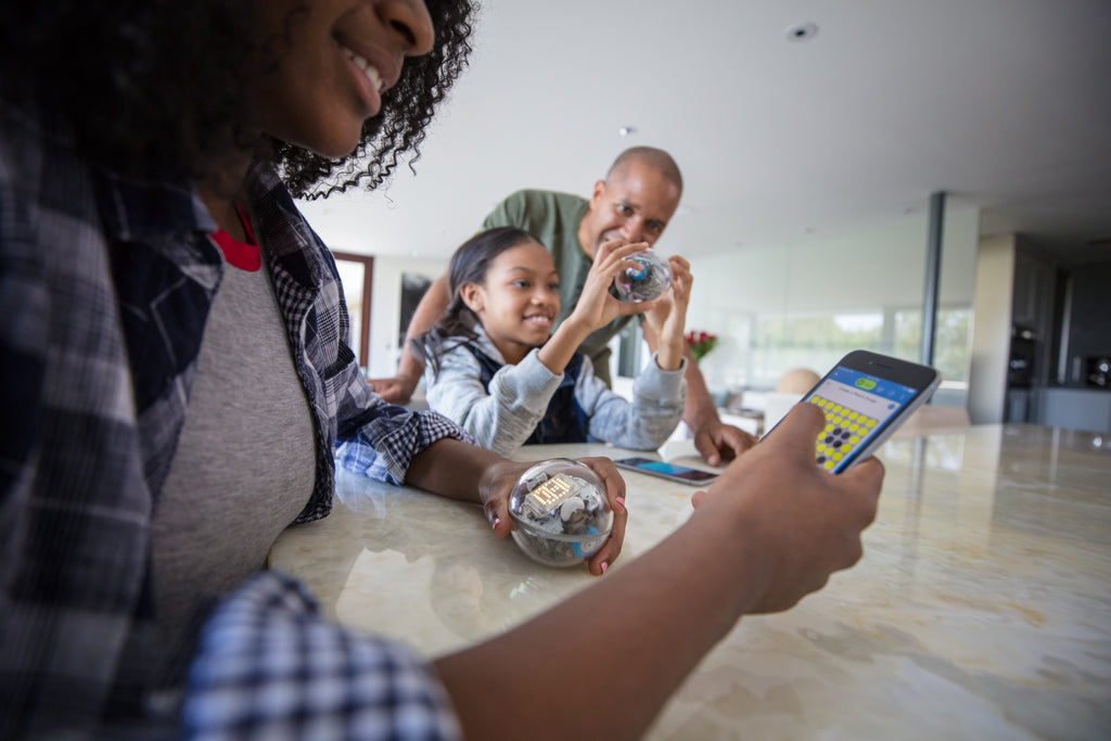 A girl and her parents sit at the kitchen counter programming a Sphero BOLT with a smartphone.