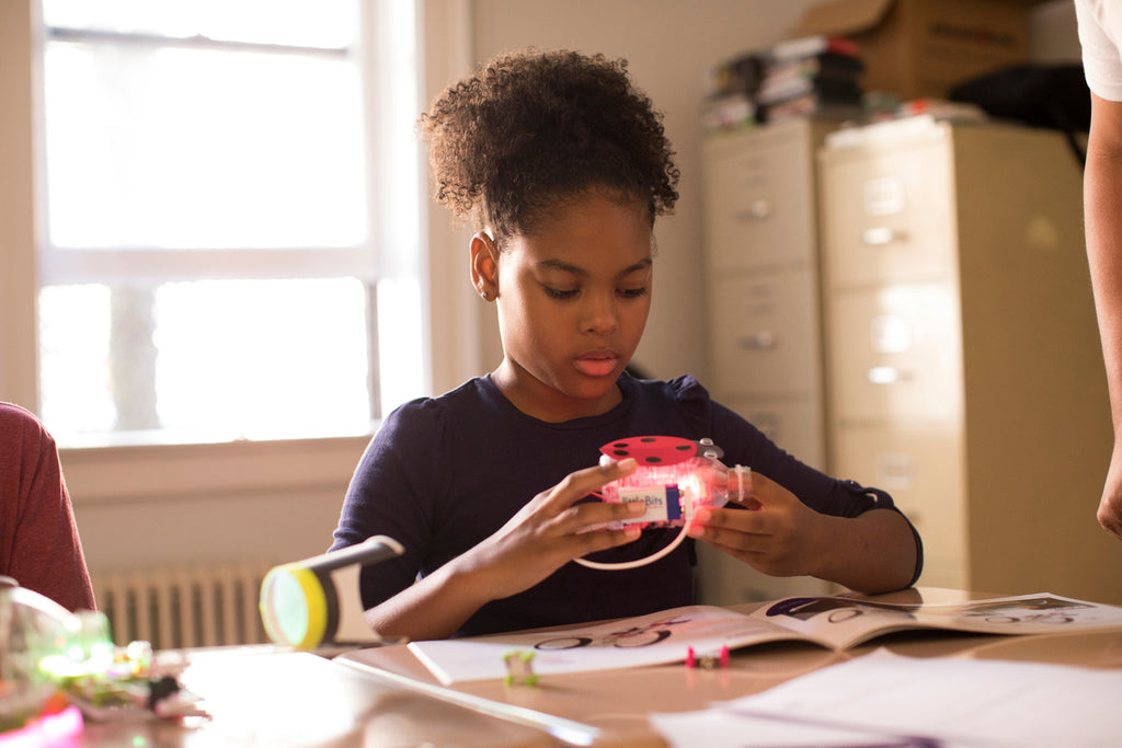 A girl sits at her desk building an invention out of littleBits and craft supplies.
